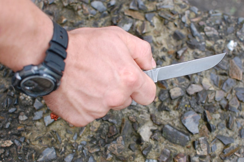 May include: A person's hand holding a silver knife with a black handle. The knife is sharp and has a pointed tip. The hand is wearing a black watch with a digital display.