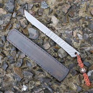 May include: A small, silver knife with a patterned blade and a brown leather sheath. The knife has a red cord with a brown bead attached to the handle.