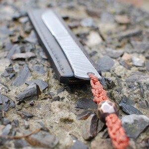 May include: A silver bookmark with a leather strap and a red braided cord with a bead. The bookmark is lying on a bed of small gray rocks.