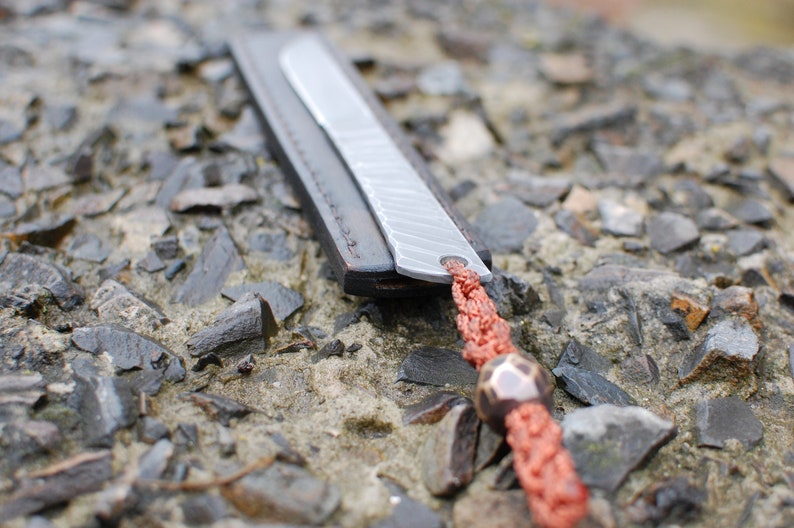 May include: A silver bookmark with a leather strap and a red braided cord with a bead. The bookmark is lying on a bed of small gray rocks.