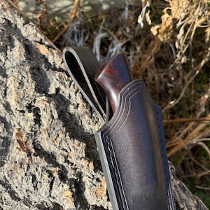 May include: A dark brown leather sheath with a stitched design and a brown wooden handle. The sheath is designed to hold a knife.