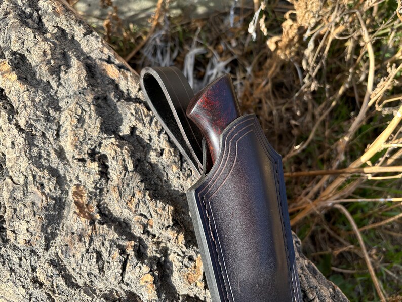 May include: A dark brown leather sheath with a stitched design and a brown wooden handle. The sheath is designed to hold a knife.