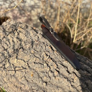 May include: A brown leather knife sheath with a metal snap closure. The sheath is resting on a piece of tree bark.