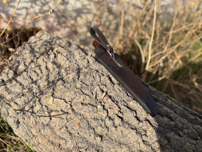 May include: A brown leather knife sheath with a metal snap closure. The sheath is resting on a piece of tree bark.