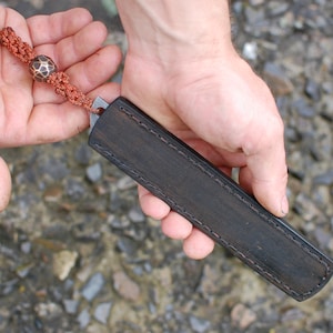 May include: A black leather knife sheath with a decorative stitch pattern. The sheath has a brown cord with a decorative bead attached.