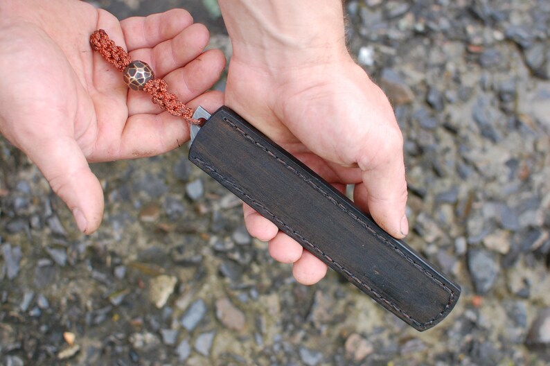 May include: A black leather knife sheath with a decorative stitch pattern. The sheath has a brown cord with a decorative bead attached.