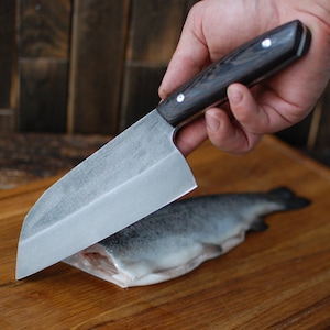 May include: A hand holding a black handled chef's knife with a silver blade, slicing a raw fish on a wooden chopping board.