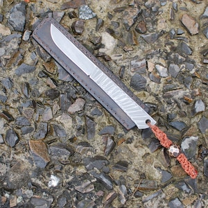 May include: A small, silver knife with a leather sheath and a red cord with a metal bead. The knife is lying on a bed of small, gray rocks.