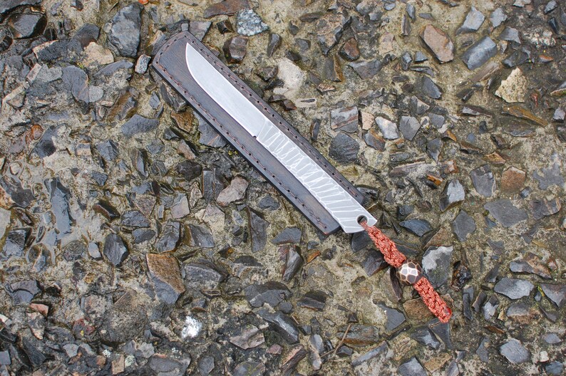 May include: A small, silver knife with a leather sheath and a red cord with a metal bead. The knife is lying on a bed of small, gray rocks.