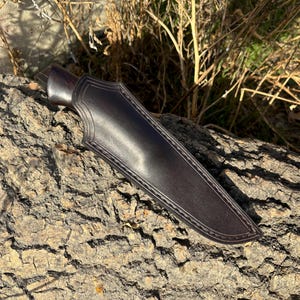 May include: A dark brown leather knife sheath with a stitched edge. The sheath is lying on a rough, brown bark surface.