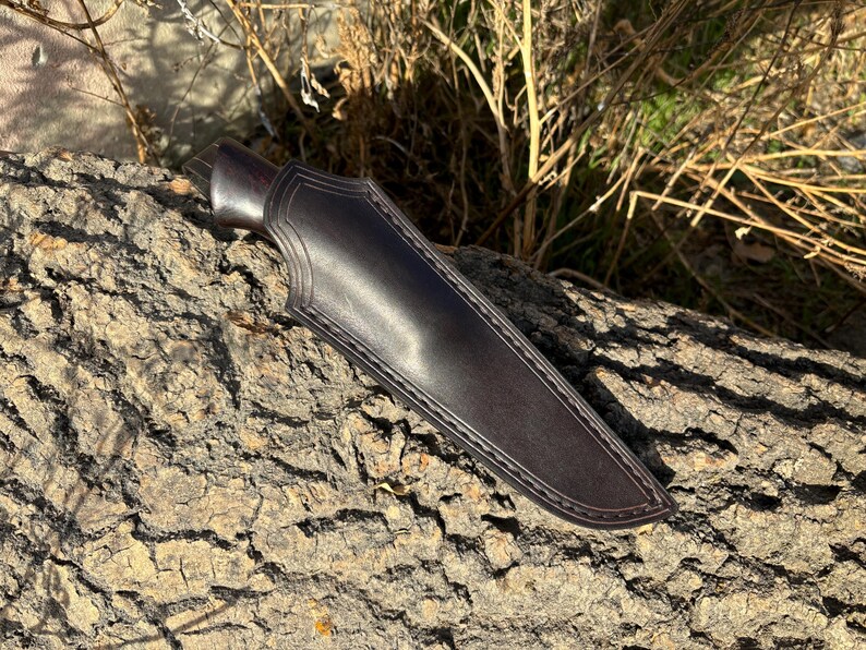 May include: A dark brown leather knife sheath with a stitched edge. The sheath is lying on a rough, brown bark surface.