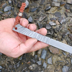 May include: A small, silver knife with a textured blade and a hole near the handle. The knife is attached to a brown cord with a copper bead.