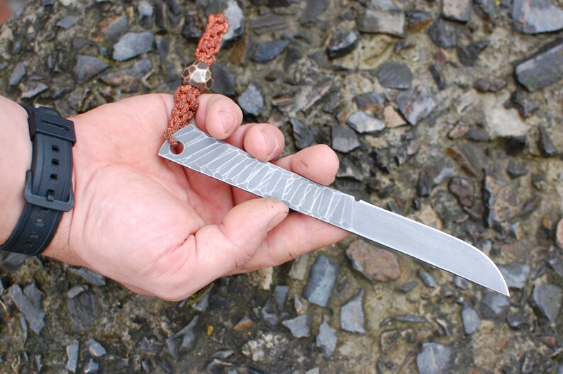 May include: A small, silver knife with a textured blade and a hole near the handle. The knife is attached to a brown cord with a copper bead.