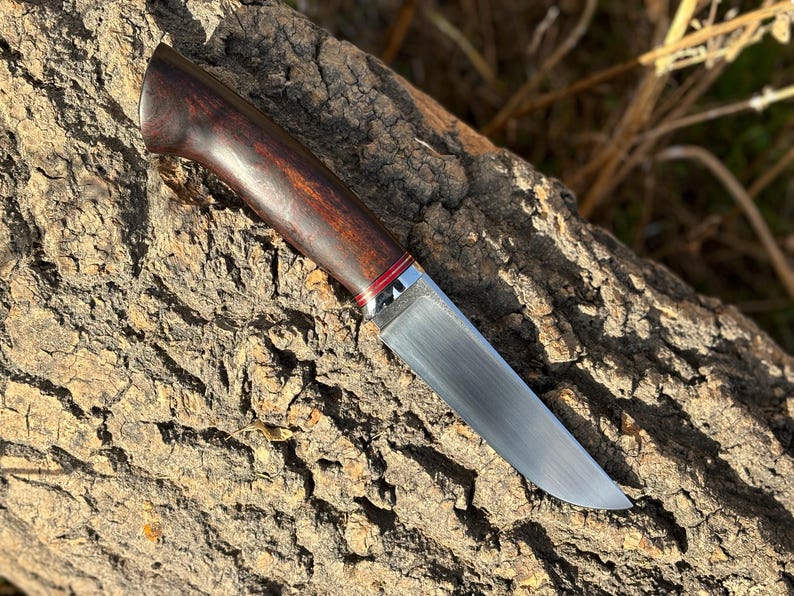 May include: A hunting knife with a dark wood handle and a silver metal bolster. The blade is made of stainless steel and has a satin finish.
