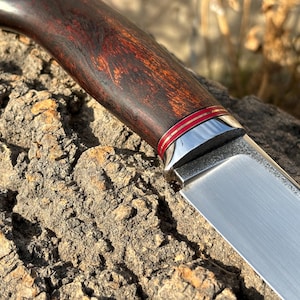 May include: A close-up of a hunting knife with a dark wood handle and a silver blade. The handle has a red and gold inlay.