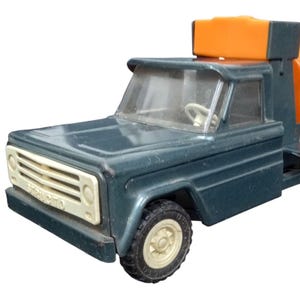 May include: A vintage toy cement mixer truck in teal and orange. The truck features a teal cab with a white grill and a rotating orange cement mixer. The wheels are black with cream-colored rims.