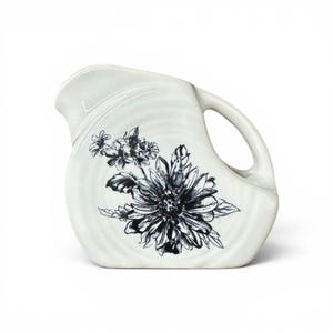 Può includere: Una brocca in ceramica color crema con un'illustrazione floreale in bianco e nero sul davanti. La brocca presenta un manico curvo, un beccuccio largo e una finitura liscia e lucida con sottili texture a cerchi concentrici sul corpo.