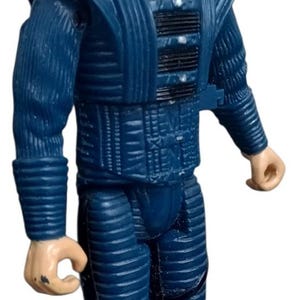 May include: A vintage action figure of a man in a dark blue futuristic uniform. The figure has orange hair and a stern expression. The uniform features ribbed detailing and black boots, typical of a science fiction theme.