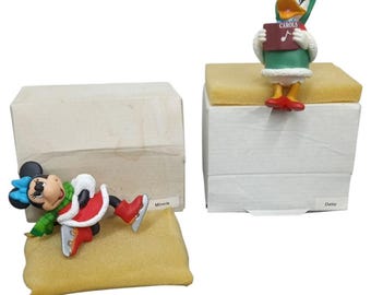 May include: Two Christmas-themed Disney figurines. One is Minnie Mouse in a red dress, green scarf, and ice skates. The other is Daisy Duck holding a sign that says "Carols". Both figurines are on top of white boxes.