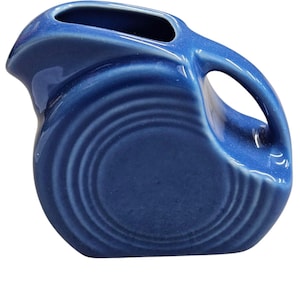 Può includere: Una brocca in ceramica blu cobalto con un manico curvo e un beccuccio. La brocca ha un corpo arrotondato con dettagli a cerchi concentrici. Il manico è a forma di anello. La brocca è di colore pieno con finitura lucida.