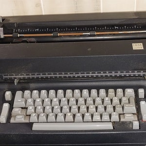 Puede incluir: Una máquina de escribir IBM Selectric II negra vintage con un teclado QWERTY completo y teclas grises. La máquina de escribir tiene una palanca de retorno de carro y un rodillo de alimentación de papel. El logotipo de IBM es visible en la parte superior derecha.