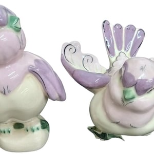 May include: Two ceramic bird figurines in a soft color palette. One bird is upright, the other has its wings spread. The birds feature lavender, white, and green accents. The figurines have a glossy finish and are likely decorative items.