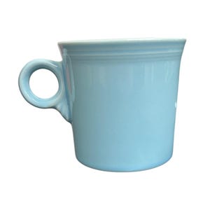Könnte beinhalten: Eine hellblaue Keramiktasse mit rundem Henkel. Die Tasse hat eine glatte, glänzende Oberfläche und einen leicht ausgestellten Rand. Die Tasse ist einfarbig hellblau.