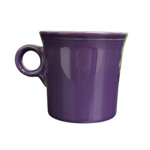 May include: A solid purple ceramic mug with a rounded handle. The mug has a slightly flared shape and a smooth, glossy finish. The handle is circular and attached to the side of the mug. The mug is designed for hot beverages.