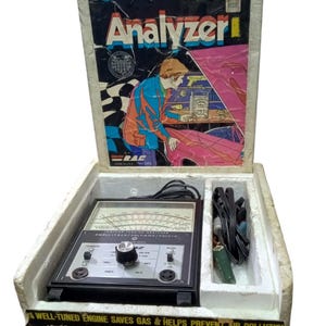 Puede incluir: Un analizador de puesta a punto de encendido negro y plateado en su caja original. La caja tiene una ilustración colorida de un automóvil y una persona trabajando en él. El analizador tiene un dial, perillas y una variedad de conectores. La caja dice "Analizador de puesta a punto de encendido" y "Un motor bien afinado ahorra gasolina y ayuda a prevenir la contaminación del aire".