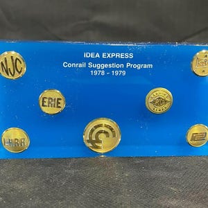 Könnte beinhalten: Eine blaue Acrylplatte mit goldenen Metallmedaillons, die Eisenbahnlogos zeigen. Die Platte trägt die Aufschrift "Idea Express Conrail Suggestion Program 1978 - 1979".