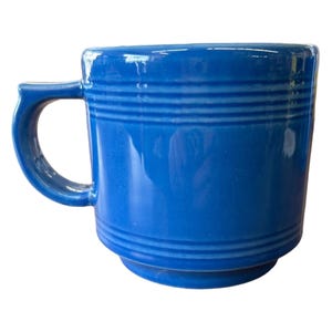 May include: A cobalt blue ceramic mug with a rounded handle. The mug features three horizontal, raised bands around the body. The mug has a glossy finish and is set against a white background.