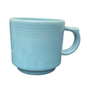 May include: A light blue ceramic mug with a rounded handle. The mug has a cylindrical shape with horizontal ridges near the top and bottom. The mug is designed to hold hot or cold beverages.