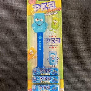 May include: A blue and yellow Pez dispenser featuring a cartoon character with a blue face and yellow eyes. The dispenser comes with three packs of blue raspberry Pez candy. The packaging says "Pez Sours" and "Exclusive Product". Total candy weight is 0.87 ounces (24.7 grams).