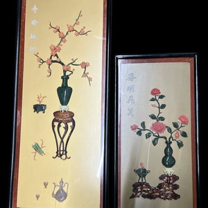 May include: Two framed pieces of Asian art with floral motifs. The left frame features a vase with a pink blossom tree and the right frame features a vase with pink roses. Both frames have gold backgrounds and black frames.