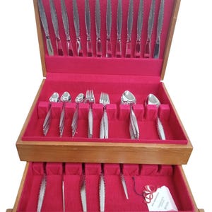 May include: A wooden flatware chest with a vibrant red velvet interior, containing a set of stainless steel utensils including knives, forks, and spoons. The chest features a pull-out drawer and a small white tag with the text Community Stainless.