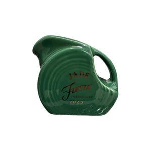 Può includere: Brocca Fiesta verde giada con beccuccio curvo e manico. La brocca ha un design a coste e le parole "JADE Fiesta INTRODUCED 2023" sono stampate in lettere dorate.