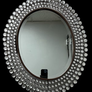 May include: Oval mirror with a dark brown frame and a border of clear, round crystals. The crystals vary in size, creating a textured, decorative edge. The mirror reflects a room with white walls and a black background.