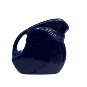 Può includere: Una brocca in ceramica blu cobalto con un manico curvo e un beccuccio. La brocca ha una finitura lucida e un corpo arrotondato. Il design è semplice ed elegante, adatta per servire bevande o come elemento decorativo.