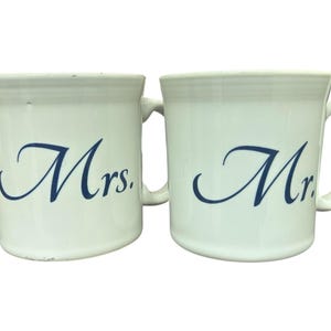 Puede incluir: Dos tazas de cerámica blancas con texto azul en cursiva. Una taza dice "Mrs." y la otra "Mr.". Ambas tazas tienen un asa en el lado derecho. Las tazas son probablemente para una pareja.