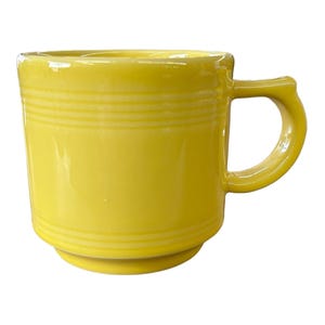 Könnte beinhalten: Eine leuchtend gelbe Keramiktasse mit rundem Boden und gebogenem Henkel. Die Tasse hat horizontale geriffelte Details um den Körper. Die Tasse hat eine einfarbige, leuchtend gelbe Farbe.