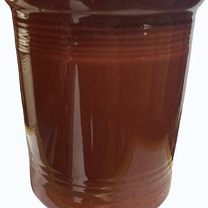 May include: A glossy, reddish-brown ceramic tumbler. The cylindrical cup features horizontal ridged detailing around the middle and a slightly flared rim. The interior is a darker shade of the same color.