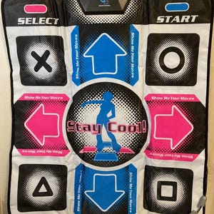 May include: A rectangular dance mat featuring a black and white graphic design with pink and blue directional arrows. The central circle displays a silhouette of a person and the text Stay Cool!. It includes Select and Start buttons.