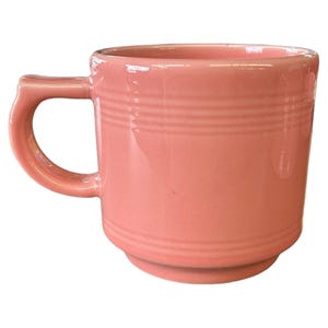 May include: A solid pink ceramic mug with a rounded handle. The mug has a stepped base and horizontal ridged detailing around the body. The mug is a light, pastel pink color.