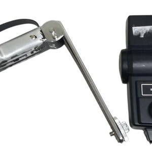 Puede incluir: Un flash Vivitar auto thyristor 283 vintage con un soporte plateado y un asa negra. El flash es negro con un panel de control y un soporte plateado. El soporte está unido a un brazo de metal plateado.
