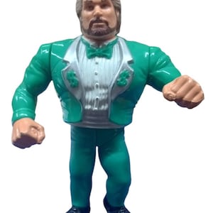 May include: An action figure of a wrestler wearing a green tuxedo with dollar sign accents. The figure has a beard and brown hair.
