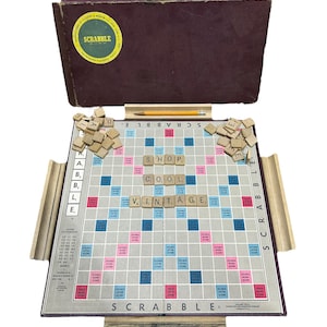 May include: A vintage Scrabble board game with wooden tiles and a brown cardboard box. The board has a light gray background with pink and blue squares. The tiles are arranged to spell out the words "SHOP", "COOL", and "VINTAGE".