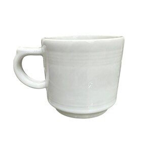 Könnte beinhalten: Eine weiße Keramiktasse mit gebogenem Henkel. Die Tasse hat ein schlichtes, klassisches Design mit leicht ausgestelltem Boden und glatter, glänzender Oberfläche. Die Tasse eignet sich für Kaffee, Tee oder andere Getränke.