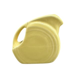 Può includere: Una brocca in ceramica giallo pallido con un manico curvo e un beccuccio. La brocca ha un corpo arrotondato con una serie di creste concentriche. La brocca è probabilmente vintage e adatta per servire bevande.