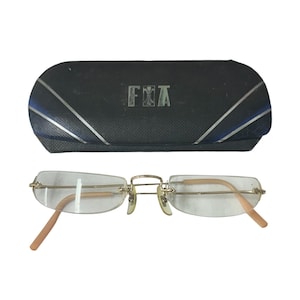 May include: Rimless eyeglasses with a gold-tone bridge and temples, paired with a black case. The case has a silver and blue stripe design and the letters "FXA" are visible on the case.