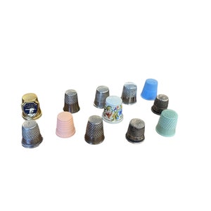 May include: A collection of 11 thimbles in various colors and designs, including silver, gold, pink, blue, and white. Some thimbles have a floral pattern, while others have a simple design.
